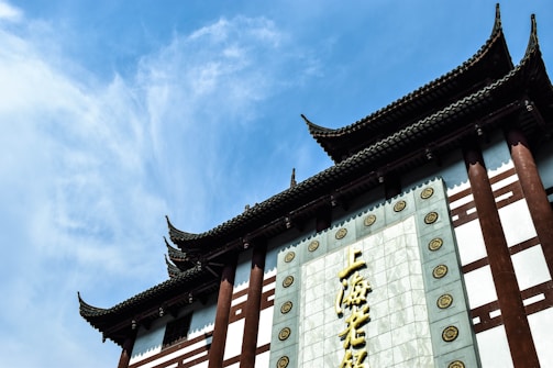 A traditional Asian architectural structure with ornate, curved roofs typical of ancient Chinese design. The building features a blend of dark wooden elements and white stone-like materials, with decorative circular motifs along a central vertical panel. Large Chinese characters are prominently displayed.
