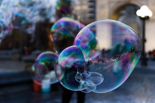 Large, colorful bubbles float in the air against a blurred urban background. The bubbles display iridescent hues, reflecting light in a spectrum of colors. There is a sense of motion and playfulness captured in the image.