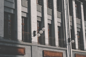 A facade of a building featuring vertical columns of white brick and gray panels. The windows have dark frames and are set within the wall structure. There are black metal railings and decorative street lamps with spherical white bulbs extending from the building.