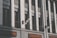 A facade of a building featuring vertical columns of white brick and gray panels. The windows have dark frames and are set within the wall structure. There are black metal railings and decorative street lamps with spherical white bulbs extending from the building.