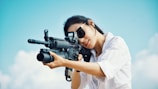 A person wearing sunglasses and a white shirt is aiming through the sight of a rifle against a bright blue sky with some clouds.