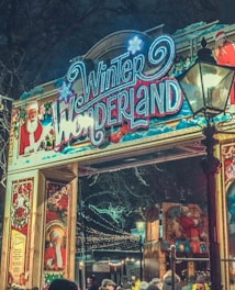 An entrance archway decorated for the Winter Wonderland event, featuring colorful lights and festive imagery, including a large sign with the words 'Winter Wonderland' in bright, illuminated script. Santa Claus imagery is prominently displayed, with depictions of Santa and festive decorations in the background. A vintage-style street lamp is visible, and a crowd of people in winter clothing gathers beneath the arch.
