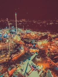 Aerial view of a vibrant nighttime amusement park with numerous rides and attractions illuminated with colorful lights. The scene includes a large circus tent near the center, several brightly lit rides, and a bustling crowd of people enjoying the festive atmosphere. In the background, a city skyline is visible against the night sky.