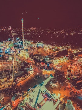 Aerial view of a vibrant nighttime amusement park with numerous rides and attractions illuminated with colorful lights. The scene includes a large circus tent near the center, several brightly lit rides, and a bustling crowd of people enjoying the festive atmosphere. In the background, a city skyline is visible against the night sky.
