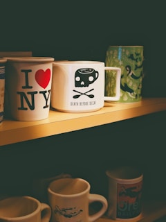 A collection of coffee mugs displayed on a shelf. The prominent mugs include one with 'I ❤ NY' in bold letters, another with a skull and crossbones design labeled 'Death Before Decaf', and a green mug featuring a mermaid pattern. The lighting casts a warm, cozy ambiance over the scene.