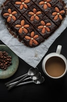 A neatly arranged brownie topped with whole almonds, forming a flower pattern on each square piece, sits on parchment paper. Nearby are a ceramic plate filled with coffee beans, four silver spoons, and a mug of coffee with a creamy texture.