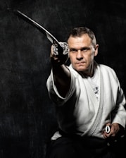 A person in a martial arts uniform is holding a katana sword with a focused expression, aiming it forward against a dark textured background. The scene conveys a sense of concentration and mastery in martial arts.