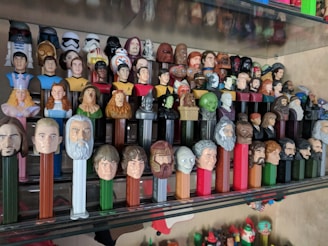 A collection of PEZ dispensers is displayed on a clear shelf, featuring a variety of character heads from popular culture, including superheroes, science fiction figures, and other recognizable characters.