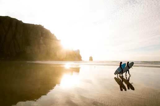 two people carrying surfboards while walking on seashore