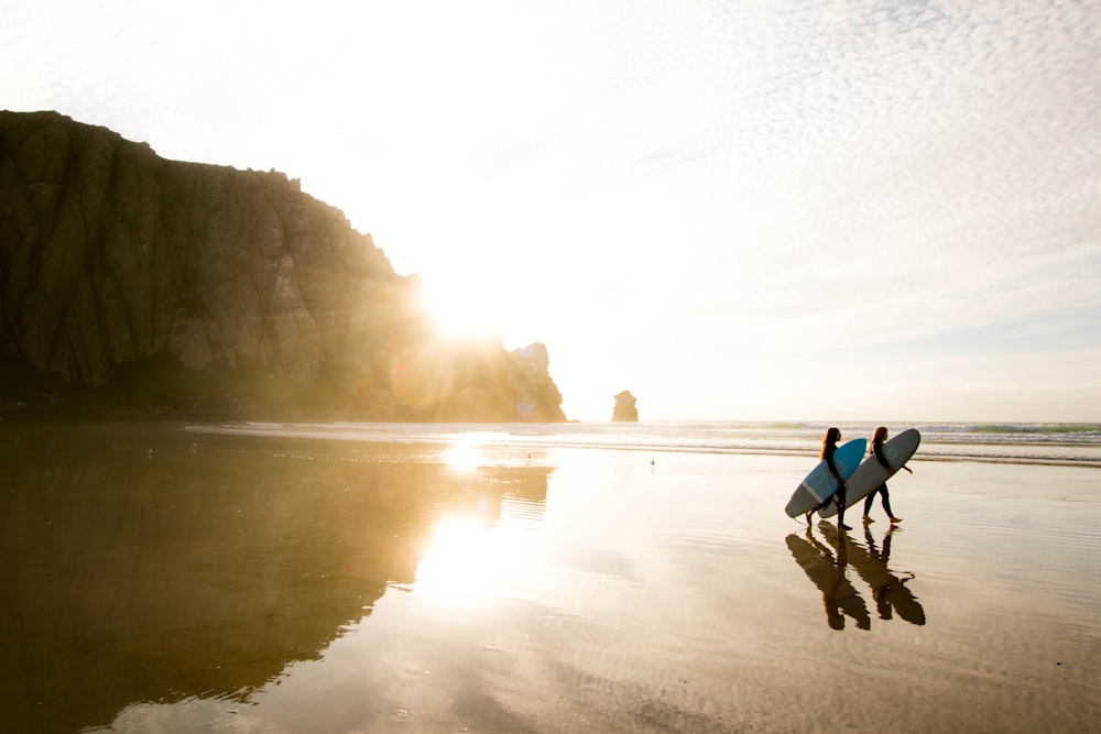 two people carrying surfboards while walking on seashore