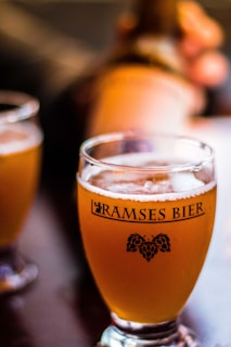 A glass of light amber beer sits on a table, filled with a frothy head. The glass is labeled 'Ramses Bier' with a small graphic of hops beneath the text. Another glass of beer is slightly blurred in the background, accompanied by a hand holding a bottle.