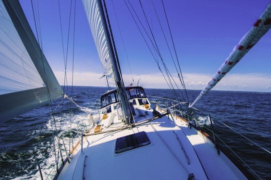 A sailing boat is navigating through a vast ocean, with its sails fully extended under a clear, vibrant blue sky. The deck is visible from a forward angle, showcasing parts of the mast and sail ropes.