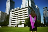 A graduate stands on a grassy area facing a group of tall modern buildings on a clear day, wearing a black gown with a purple sash. The scene conveys a sense of accomplishment and anticipation.