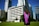A graduate stands on a grassy area facing a group of tall modern buildings on a clear day, wearing a black gown with a purple sash. The scene conveys a sense of accomplishment and anticipation.