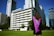 A graduate stands on a grassy area facing a group of tall modern buildings on a clear day, wearing a black gown with a purple sash. The scene conveys a sense of accomplishment and anticipation.
