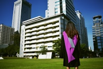 A graduate stands on a grassy area facing a group of tall modern buildings on a clear day, wearing a black gown with a purple sash. The scene conveys a sense of accomplishment and anticipation.