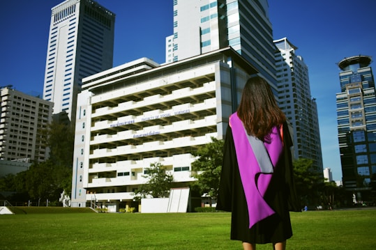 A graduate stands on a grassy area facing a group of tall modern buildings on a clear day, wearing a black gown with a purple sash. The scene conveys a sense of accomplishment and anticipation.