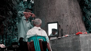 man sitting on green chair facing mirror while the other man standing cutting's man hair at daytime