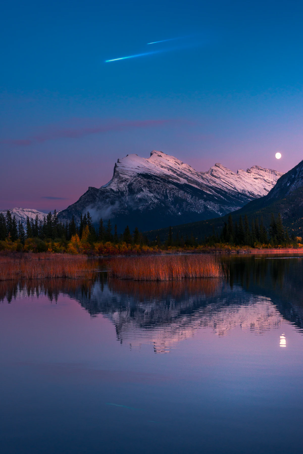 A snowy peak and a full moon reflected in Vermilion Lakes outside Banff at dusk