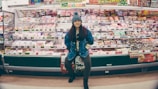 A person stands confidently in front of a well-stocked supermarket shelf filled with various packaged food items. The individual is wearing a denim jacket and a knit hat, appearing casual and relaxed. The shelves are colorful, with different products neatly arranged and price tags visible.