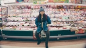 A person stands confidently in front of a well-stocked supermarket shelf filled with various packaged food items. The individual is wearing a denim jacket and a knit hat, appearing casual and relaxed. The shelves are colorful, with different products neatly arranged and price tags visible.