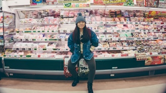 A person stands confidently in front of a well-stocked supermarket shelf filled with various packaged food items. The individual is wearing a denim jacket and a knit hat, appearing casual and relaxed. The shelves are colorful, with different products neatly arranged and price tags visible.