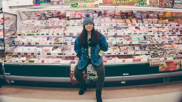 A person stands confidently in front of a well-stocked supermarket shelf filled with various packaged food items. The individual is wearing a denim jacket and a knit hat, appearing casual and relaxed. The shelves are colorful, with different products neatly arranged and price tags visible.