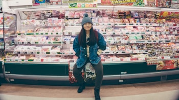 A person stands confidently in front of a well-stocked supermarket shelf filled with various packaged food items. The individual is wearing a denim jacket and a knit hat, appearing casual and relaxed. The shelves are colorful, with different products neatly arranged and price tags visible.