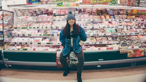 A person stands confidently in front of a well-stocked supermarket shelf filled with various packaged food items. The individual is wearing a denim jacket and a knit hat, appearing casual and relaxed. The shelves are colorful, with different products neatly arranged and price tags visible.
