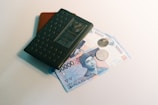 A green Indonesian passport is placed atop two banknotes of 50,000 Indonesian Rupiahs and two coins. The passport has a textured surface and the money is slightly visible beneath it.