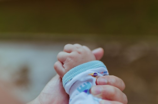 A close-up of an adult hand gently holding a baby's hand, conveying a sense of care and protection. The baby is wearing a white garment with a blue trim and colorful designs.