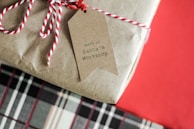 A gift wrapped in plain brown paper is tied with a red and white striped string. Attached is a brown paper tag with the text 'made in Santa's Workshop'. The background consists of a festive red color and a glimpse of a plaid pattern below the gift.