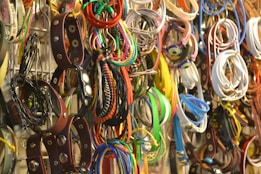 A collection of various belts and bracelets hangs in a disorganized manner on a rack, showcasing a wide array of colors and styles. There are leather items with metal studs, colorful fabric bracelets, and woven accessories.