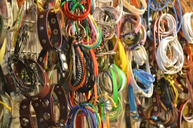 A collection of various belts and bracelets hangs in a disorganized manner on a rack, showcasing a wide array of colors and styles. There are leather items with metal studs, colorful fabric bracelets, and woven accessories.