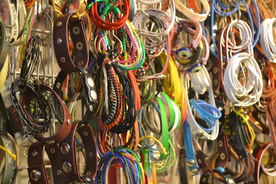 A collection of various belts and bracelets hangs in a disorganized manner on a rack, showcasing a wide array of colors and styles. There are leather items with metal studs, colorful fabric bracelets, and woven accessories.