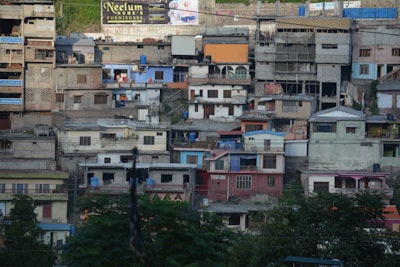 A densely packed hillside neighborhood with a variety of colorful, multi-story buildings stacked closely together. The buildings exhibit different architectural styles and a mix of materials, including brick and concrete. There are several water tanks visible on rooftops, and vegetation appears in the foreground.