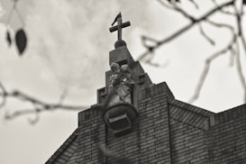 A sepia-toned image of a church steeple featuring a cross at the top and a statue of a religious figure below. The structure is made of brick and there are blurred branches in the foreground.