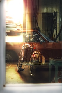 A vintage bicycle is seen indoors through a large window, with light reflections creating a dreamy atmosphere. The background features an old car partially visible, contributing to a nostalgic feel. The setting appears to be a garage or workshop, and the lighting is warm and soft.