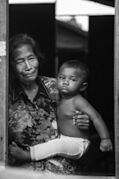 An elderly person cradles a young child while standing by a doorframe. The expression on the child's face is serious, and both figures are captured in black and white, adding a timeless and intimate feel to the scene. The elder's clothing features a floral pattern.