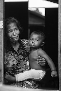An elderly person cradles a young child while standing by a doorframe. The expression on the child's face is serious, and both figures are captured in black and white, adding a timeless and intimate feel to the scene. The elder's clothing features a floral pattern.