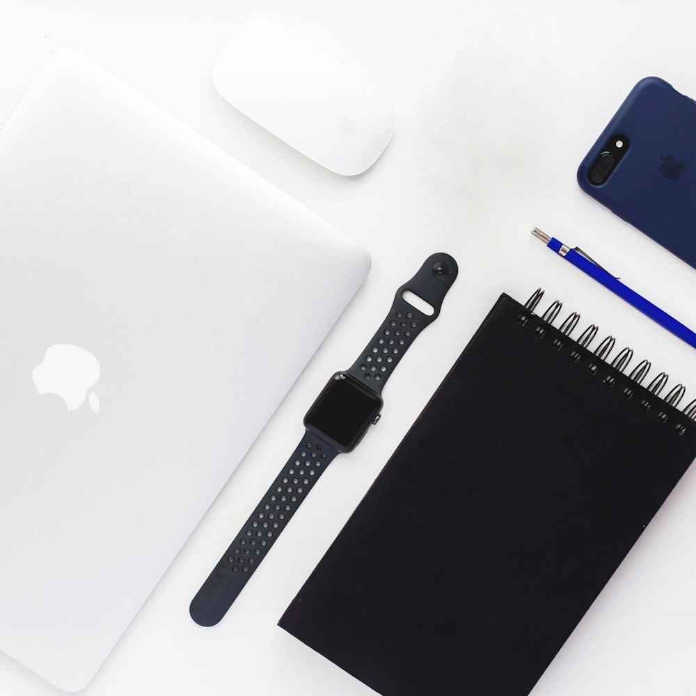 Fotografía plana de funda de aluminio gris espacial Apple Watch con correa deportiva Nike, libro en espiral, MacBook blanco, Apple Magic Mouse y iPhone 7 Plus negro con funda azul
