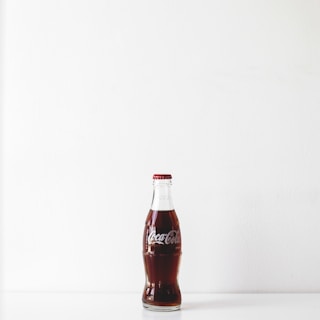A classic glass bottle of Coca-Cola sits upright on a white surface against a plain white background. The bottle is filled with dark brown liquid and sealed with a red and silver cap.