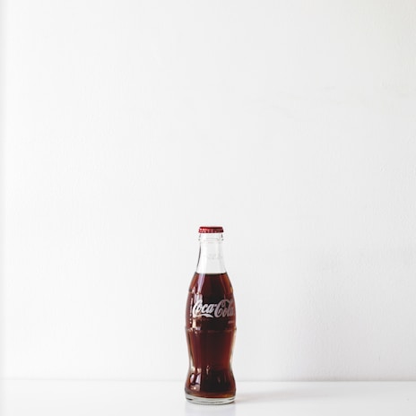 A classic glass bottle of Coca-Cola sits upright on a white surface against a plain white background. The bottle is filled with dark brown liquid and sealed with a red and silver cap.