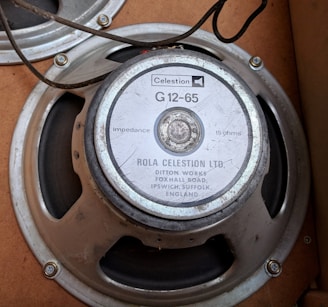 A vintage speaker from Rola Celestion Ltd. is shown, featuring a model G12-65. The speaker is mounted in a wooden enclosure, with visible electrical wiring and metal components. It has the model specifications and addresses inscribed on the backplate.