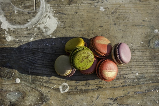 A group of colorful macarons is arranged on a rustic wooden surface, casting a shadow in the sunlight. The macarons include shades of green, yellow, red, and purple, with some displaying unique patterns and textures.