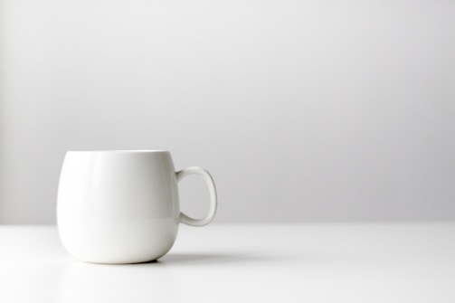 A minimalist ceramic mug in matte beige, photographed against an off-white background with diffused light.