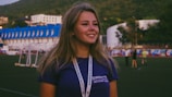 A young woman with long hair stands outdoors on a sports field, wearing a dark blue shirt and a lanyard around her neck. In the background, there are people walking and various buildings with blue roofs against a backdrop of hills.