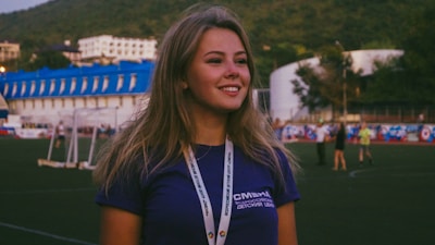 A young woman with long hair stands outdoors on a sports field, wearing a dark blue shirt and a lanyard around her neck. In the background, there are people walking and various buildings with blue roofs against a backdrop of hills.