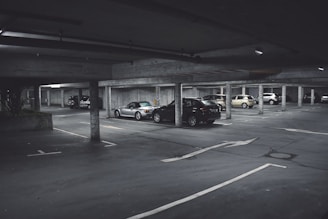 A dimly lit parking garage with several cars parked in marked spaces. Concrete pillars and overhead beams are visible, with directional arrows on the ground indicating traffic flow. The scene appears somewhat deserted with no visible people around.