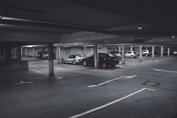 A dimly lit parking garage with several cars parked in marked spaces. Concrete pillars and overhead beams are visible, with directional arrows on the ground indicating traffic flow. The scene appears somewhat deserted with no visible people around.